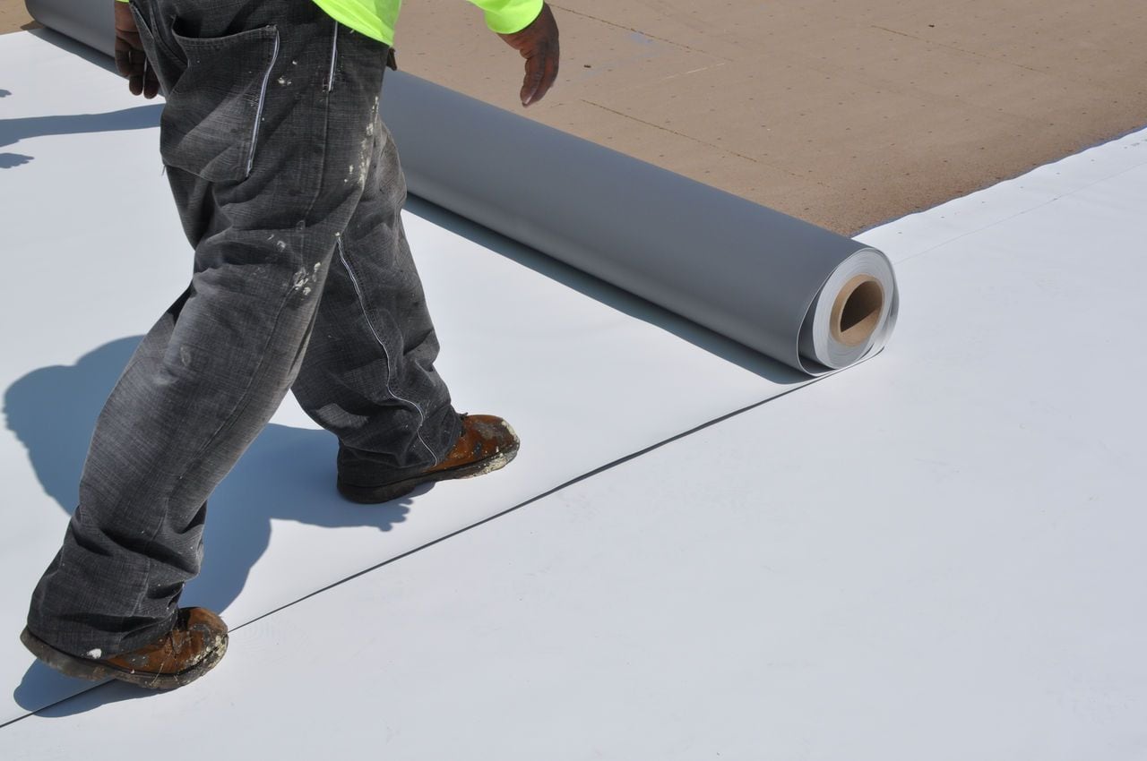 A construction worker unrolling a white single-ply roofing membrane on a commercial roof.